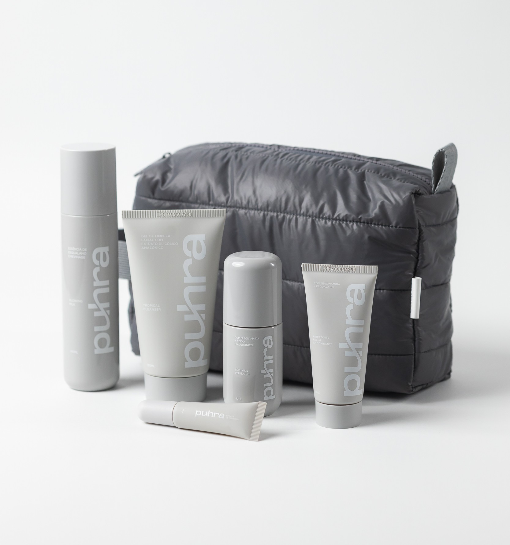 Kit de skincare Puhra composto por cinco produtos em embalagens minimalistas na cor cinza: um frasco spray, dois tubos de creme de tamanhos diferentes, um sérum roll-on e um batom/produto labial, dispostos à frente de uma nécessaire acolchoada cinza escuro com zíper